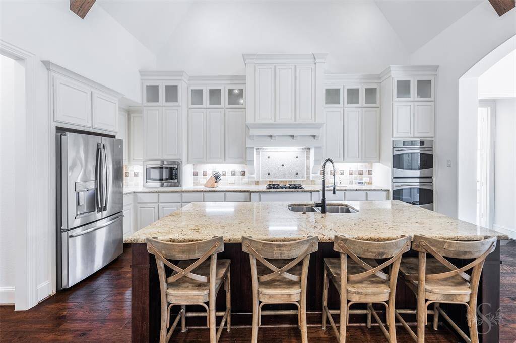 This modern kitchen features high ceilings, sleek white cabinetry, a spacious granite island with seating space, stainless steel appliances, and wood flooring, creating a bright and functional space for cooking and entertaining.