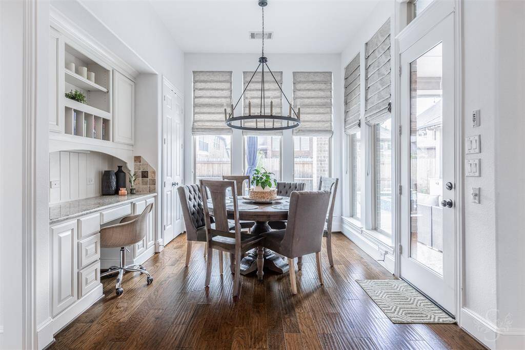 This bright breakfast room features hardwood floors, a built-in desk, and large windows with Roman shades. A modern chandelier adds elegance, and glass doors open to the outdoor space, enhancing natural light.