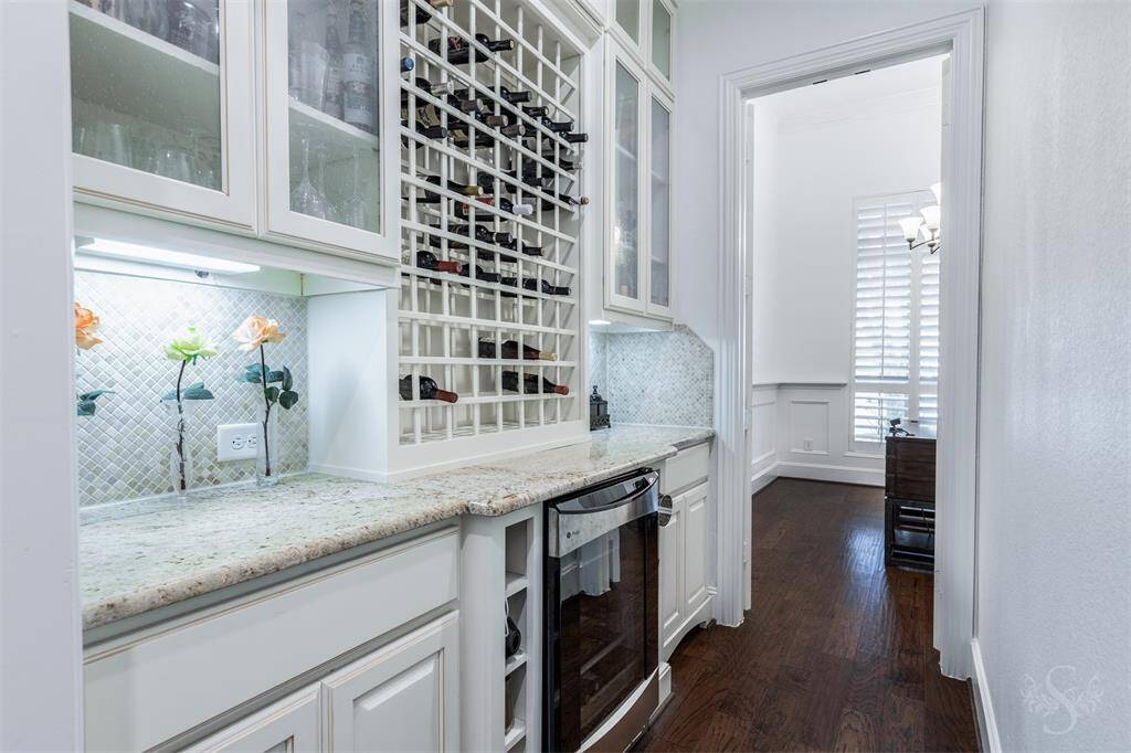 This photo showcases a stylish butler's pantry with white cabinetry, granite countertops, a beverage fridge and a built-in wine rack. The space features glass-front cabinets, under-cabinet lighting, and a mosaic tile backsplash, leading into the well-lit dining room.