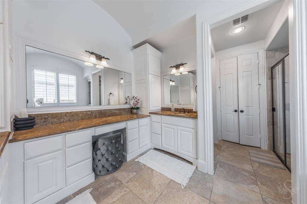 The primary bathroom features spacious dual vanities with granite countertops, ample cabinet storage, and elegant lighting. It includes a large mirror, a walk-in shower, and natural light from a nearby window, creating a bright and inviting atmosphere.