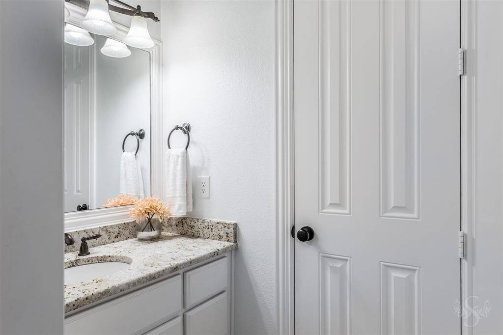 This secondary bathroom features a bright, modern design with a granite countertop, sleek fixtures, and white cabinetry. The space is accented by a large mirror and elegant lighting.