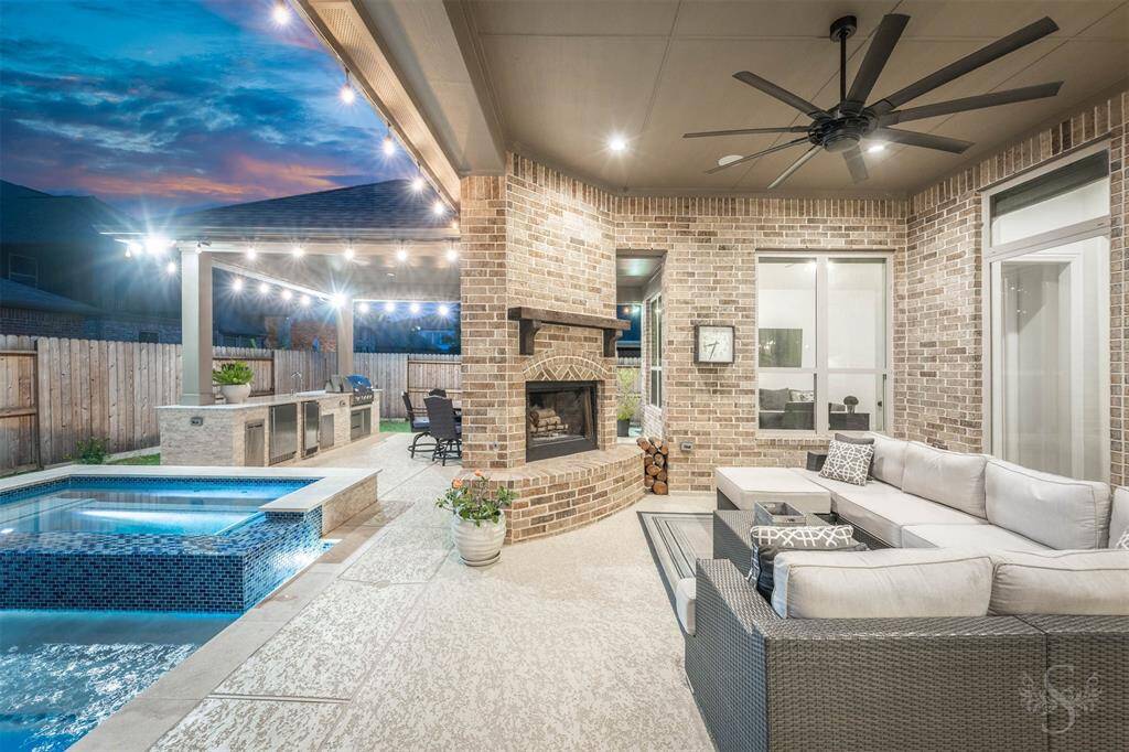 This photo showcases the luxurious outdoor living space featuring a cozy seating area with a brick fireplace, a modern ceiling fan, and elegant lighting. Adjacent is the sparkling pool and spa, alongside an outdoor kitchen with ample counter space, perfect for entertaining.