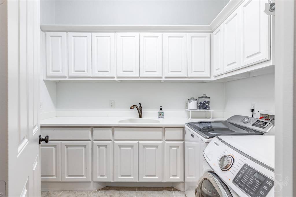 This photo showcases a bright, modern laundry room with ample white cabinetry for storage, a sleek countertop with a sink. The neutral color palette and tidy layout create a functional and inviting space.