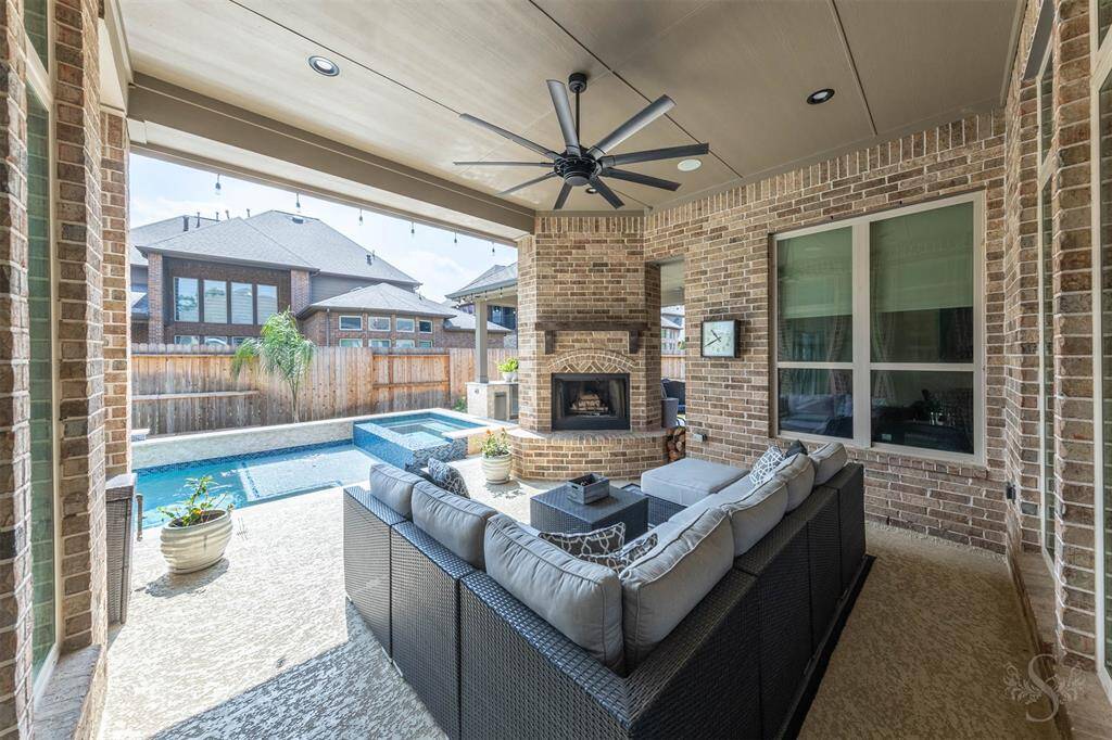 This photo showcases a stylish outdoor patio with a cozy seating area, ceiling fan, and fireplace. It overlooks the sparkling pool and spa, providing an inviting space for relaxation and entertainment. The brick architecture and covered ceiling add a touch of elegance.
