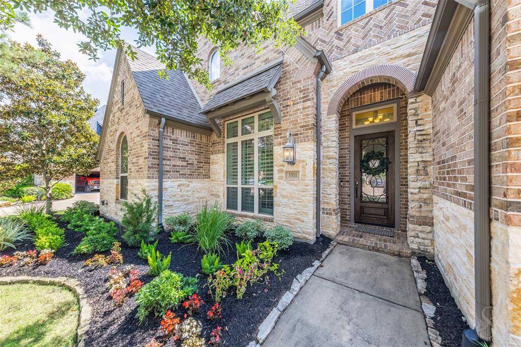 Such a charming entrance to this home with a brick and stone exterior, a welcoming front door with decorative glass, and well-maintained landscaping featuring vibrant plants and flowers.