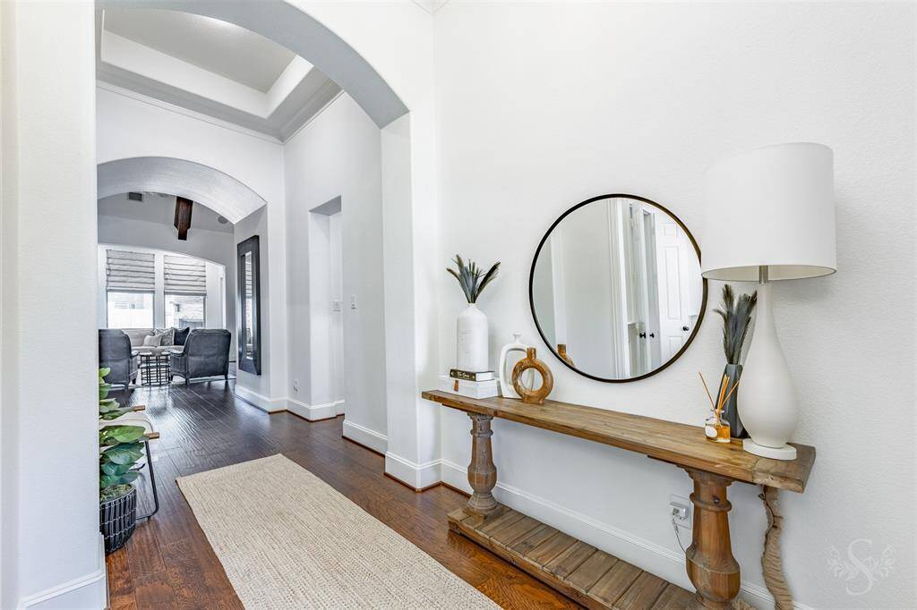 This photo showcases a bright and inviting hallway. The space features elegant archways, white walls, and warm wooden flooring leading into a well-lit living area with large windows.