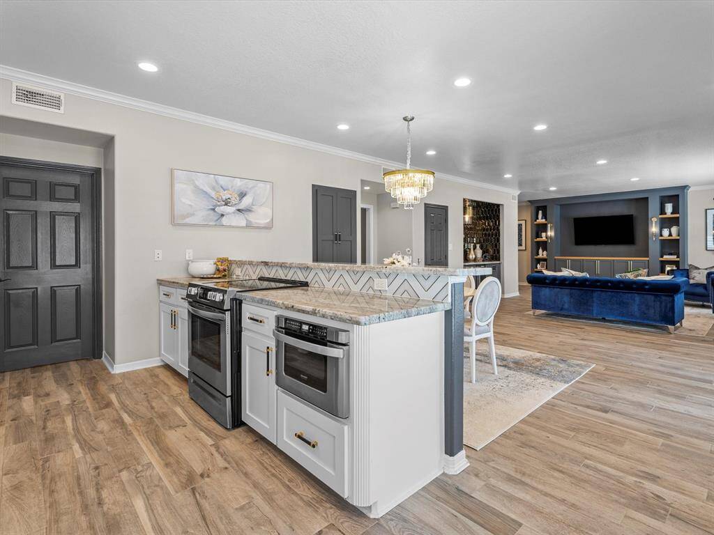 This open-concept living space features a modern kitchen with a stylish island, sleek countertops, and stainless steel appliances. The area flows into a spacious living room with elegant wood flooring, a chandelier, and a cozy entertainment setup.