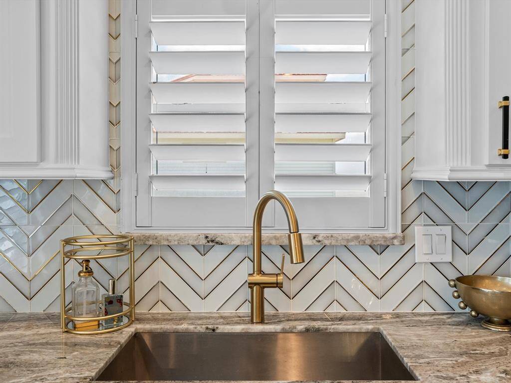 This kitchen features a modern design with a sleek brass faucet, chevron-patterned tile backsplash, and elegant marble countertops. White shutters provide natural light and privacy, while decorative accents add a touch of luxury.