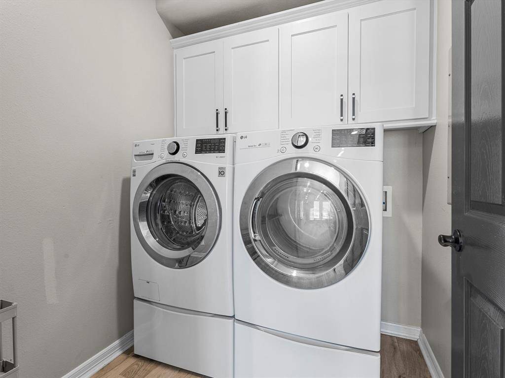 This photo shows a modern laundry room featuring a front-loading washer and dryer set. There are white cabinets above for additional storage, and the space has a clean, minimalist design with light-colored walls and flooring.