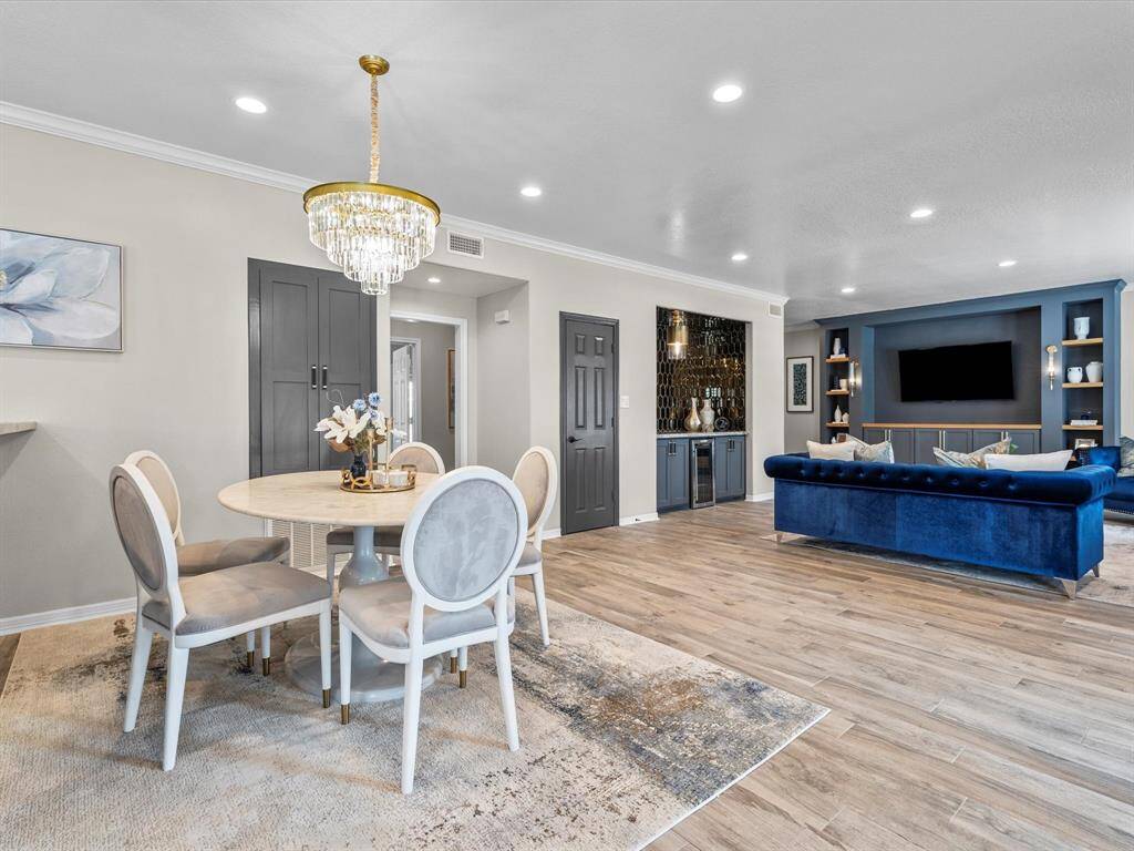 This spacious, modern living area features elegant wood flooring, a stylish blue sofa, and a chic dining space under a crystal chandelier. Built-in shelving and a sleek wet bar add functionality and sophistication, perfect for entertaining.