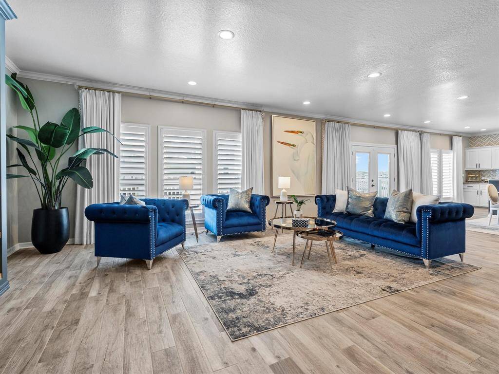 This spacious living room features elegant blue sofas, a stylish area rug, and abundant natural light from large windows with white shutters. The open layout seamlessly connects to the kitchen, creating a welcoming and modern atmosphere perfect for entertaining or relaxing.