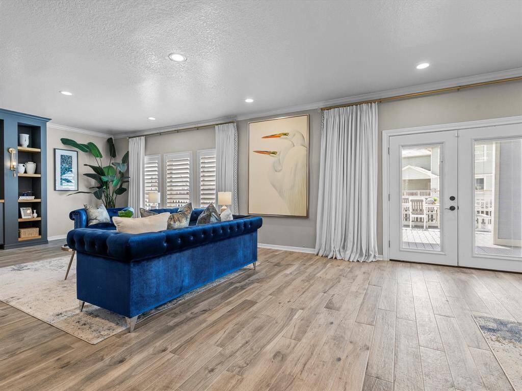 This living room features elegant hardwood floors and a cozy blue velvet sofa. It includes built-in shelving, stylish artwork, and large windows with white shutters and curtains, allowing natural light to flow in. French doors open to an outdoor area, enhancing the spacious feel.
