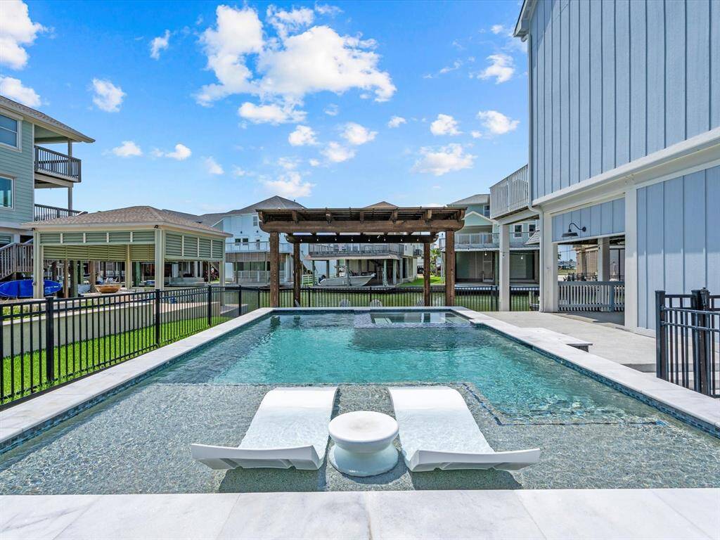 This photo showcases a modern backyard pool area with sleek lounge chairs and a small round table. The space is enclosed by a fence, surrounded by neighboring houses, and features a pergola for shade. It's a perfect spot for relaxation and entertaining.