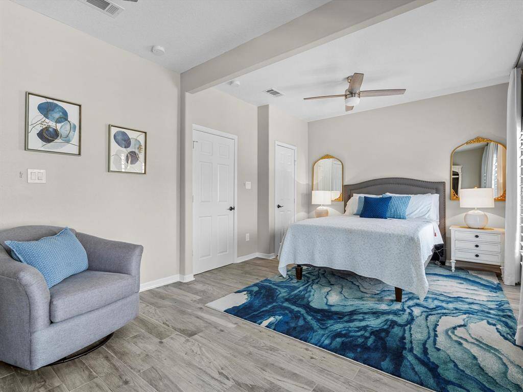 This spacious bedroom features a modern design with neutral walls and wood-like flooring. It includes a comfortable bed with plush blue accents, elegant mirrors, and a cozy armchair. The room is brightened by natural light and stylish artwork.