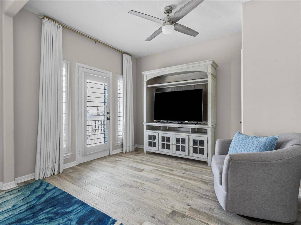 This cozy living area features a modern ceiling fan, wood-look flooring, and a large entertainment center with a TV. Natural light pours in through the glass doors with elegant white drapes, complemented by a comfortable gray chair with a blue accent pillow.
