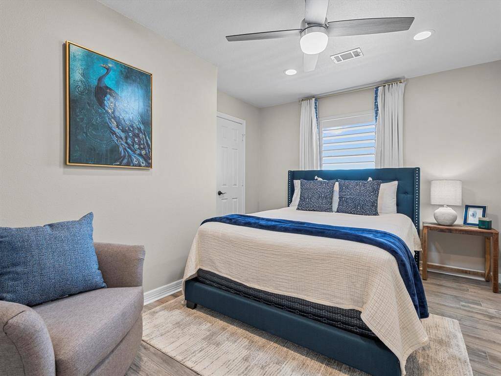 This bedroom features a cozy, modern design with a stylish bed, accent chair, and a peacock-themed artwork. It has wood flooring, a ceiling fan, and ample natural light from the window, creating a welcoming atmosphere.