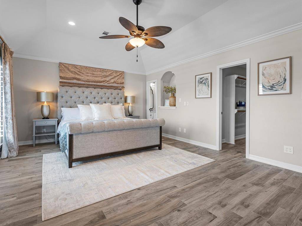 This spacious bedroom features a stylish, tufted bed with elegant bedding, modern nightstands, and a ceiling fan. The room is enhanced by wood-look flooring, soft lighting, and neutral-toned walls, with access to a walk-in closet.