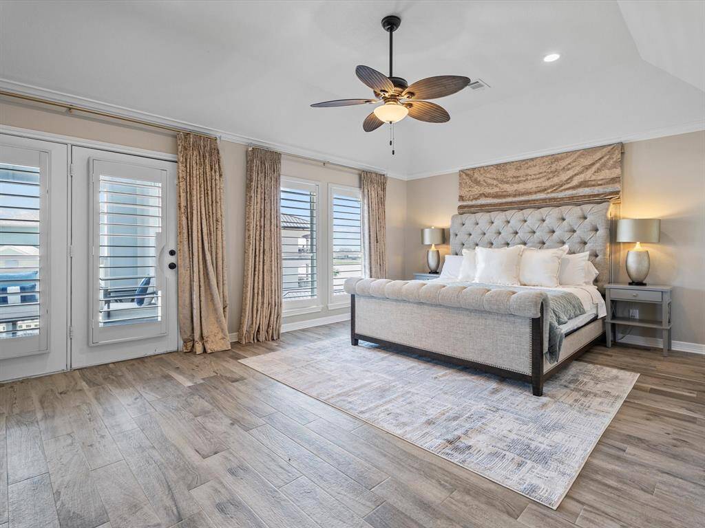 This spacious bedroom features elegant wood flooring, a plush tufted bed, and a ceiling fan. Natural light streams through large windows and French doors, enhanced by luxurious curtains, creating a bright and inviting atmosphere.