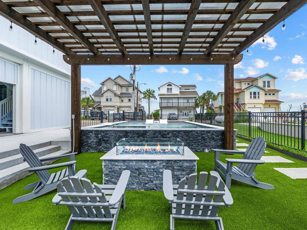 This charming backyard features a modern stone fire pit surrounded by cozy Adirondack chairs on lush artificial turf. A wooden pergola adds character, while a sleek pool and a view of neighboring houses complete this inviting outdoor space.