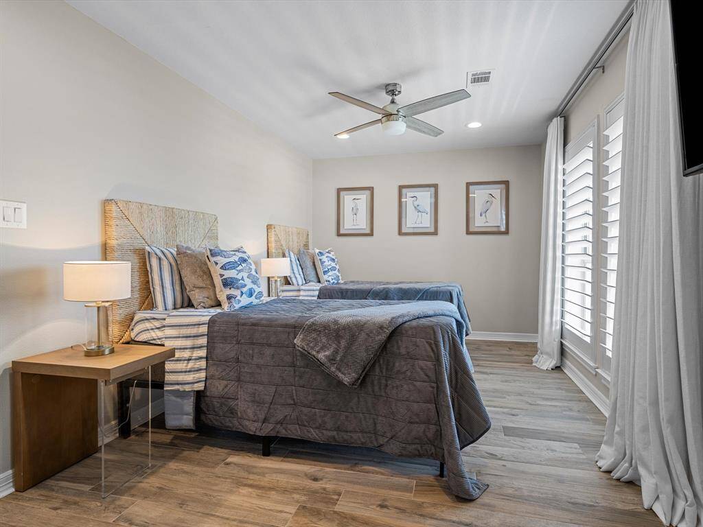 This bedroom features two cozy twin beds with stylish bedding, a modern ceiling fan, and elegant wall art. Large windows with shutters and soft curtains provide ample natural light, while the wood flooring adds warmth to the space.