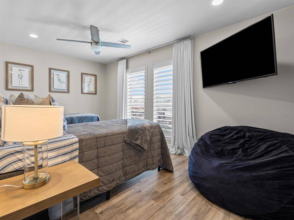 This cozy bedroom features a large bed with plush bedding, a wall-mounted TV, and ample natural light from the windows with white shutters. The room includes a ceiling fan, framed artwork, a bean bag chair, and stylish wood flooring for a modern touch.