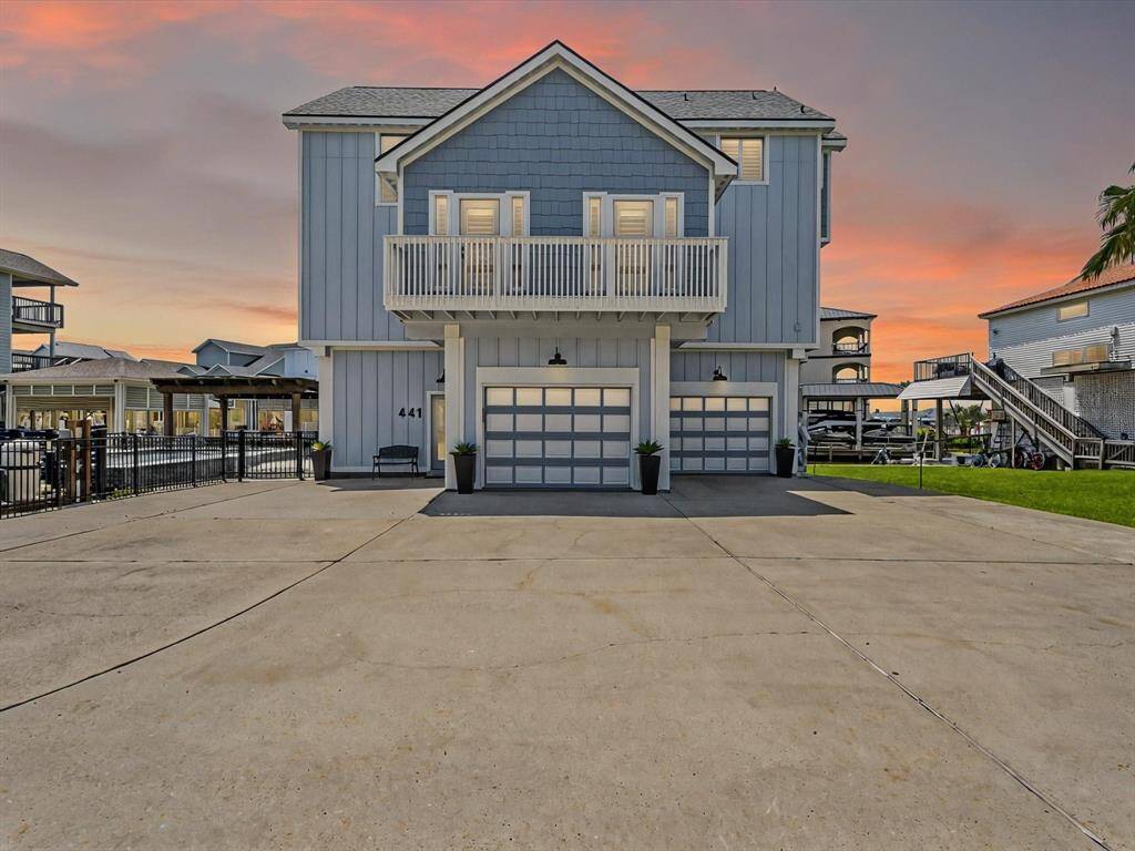 This modern coastal home features a clean, blue-gray exterior with a charming front balcony. It has a spacious driveway leading to a double garage, ideal for multiple vehicles. The property is set against a beautiful sunset backdrop, offering a serene and inviting atmosphere.
