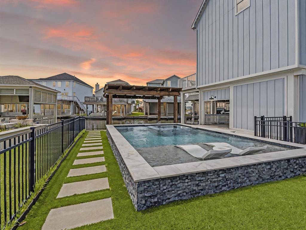 This photo showcases a modern backyard with a stylish pool surrounded by a stone border and a sleek metal fence. There's a wooden pergola for shade, and the area features well-maintained grass with stepping stones. The setting is serene, perfect for relaxation or entertaining.