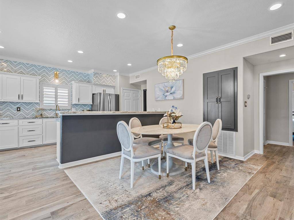 This photo showcases a modern kitchen and dining area with a chic design. The kitchen features white cabinetry, stainless steel appliances, and a stylish chevron backsplash. The dining area includes a round table with elegant chairs and a striking chandelier. The space is bright and open, with light wood flooring and contemporary decor.