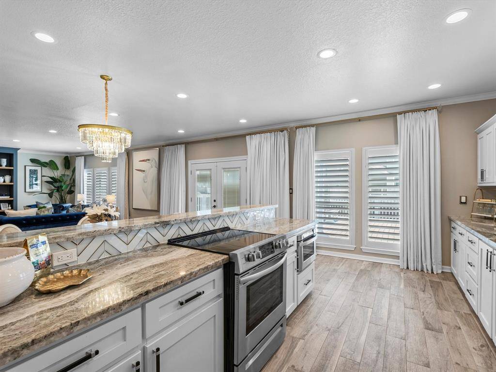 This photo showcases a modern kitchen with elegant granite countertops and a stylish backsplash. It features a large island with a built-in oven and ample cabinetry for storage. The space is bright, thanks to recessed lighting and large windows with plantation shutters. A chic chandelier adds a touch of luxury, and the open layout connects seamlessly to the living area.