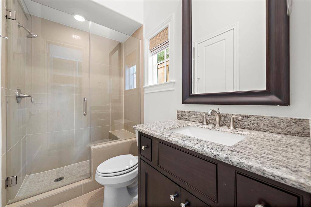 Spa-inspired secondary bath features a frameless glass walk-in shower, modern tilework, and an oversized vanity with granite countertops. Custom cabinetry, designer fixtures, and natural light through the window create a refined, comfortable retreat for guests or residents.