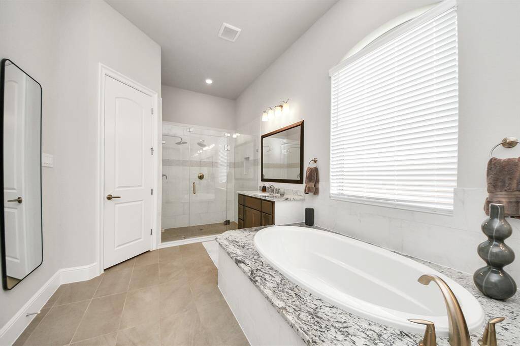 This spacious bathroom features a central soaking tub, dual vanities with granite countertops, and a large walk-in shower. The neutral color palette and modern lighting create a serene, luxurious feel.