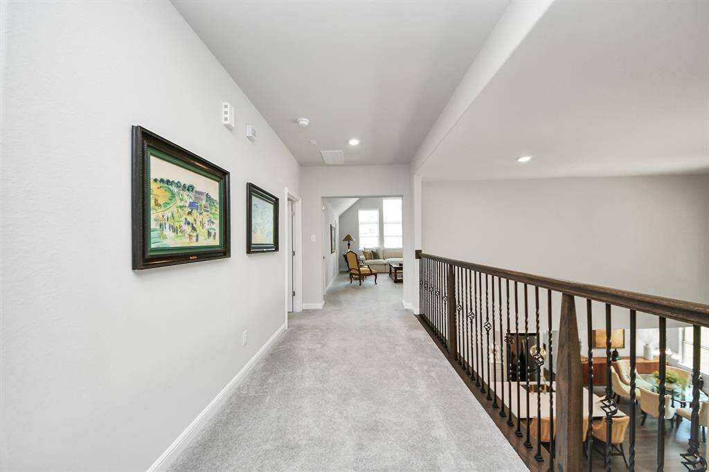 Featuring a well-lit, carpeted hallway. It leads to a cozy family room/game room with natural light. The hallway overlooks an open space with a wooden and iron railing, offering a view of the room below.