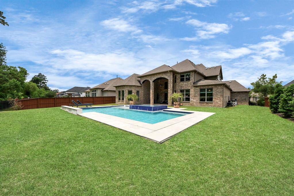 Spacious backyard featuring a modern swimming pool and spa, surrounded by lush greenery. The house has a brick exterior with covered patio areas, perfect for outdoor relaxation and entertaining.