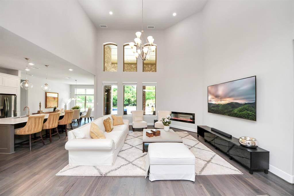 This spacious, modern living area features high ceilings, abundant natural light, and a sleek open concept. It includes a cozy sitting area with a wall-mounted TV, an elegant chandelier, and a fireplace. The adjacent kitchen boasts a large island with bar seating and pendant lighting, seamlessly connecting to the dining area. Large windows and glass doors provide views of the outdoors, enhancing the home's airy feel.
