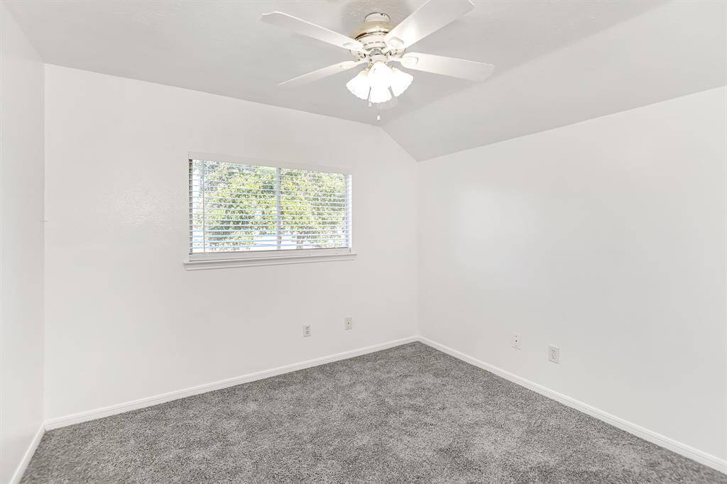 Bright and inviting room with fresh white walls, plush gray carpeting, and a ceiling fan. The window offers pleasant natural light and a view of greenery, perfect for a cozy bedroom or a home office.