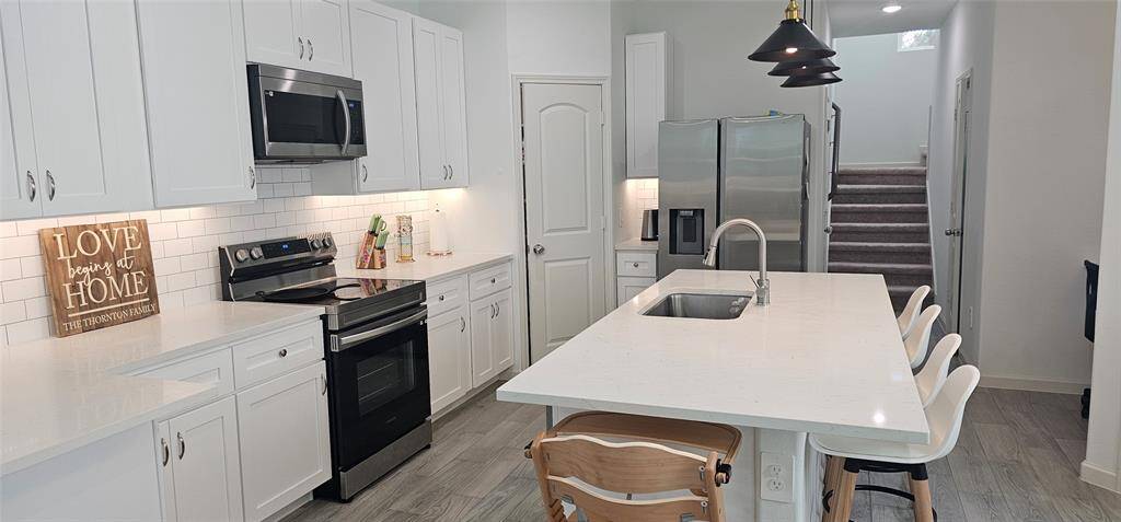 kitchen with sleek white cabinets, stainless steel appliances, and a spacious island. Features a stylish subway tile backsplash, pendant lighting, and plenty of natural light. Adjacent to a staircase leading to the upper floor.