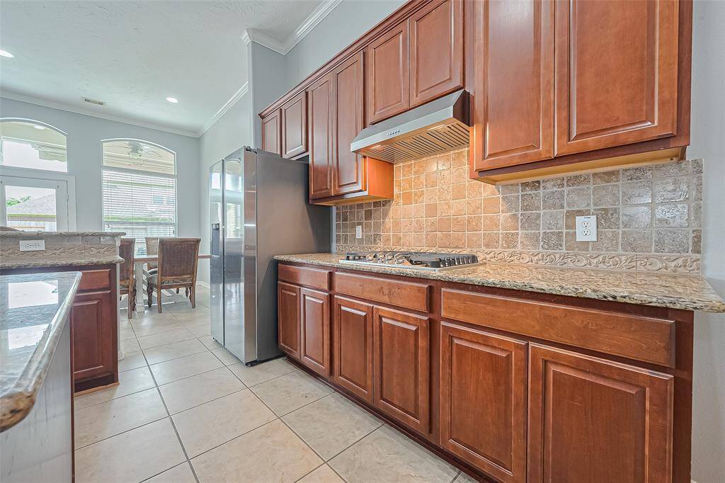 This bright kitchen features rich wooden cabinets, granite countertops, a gas cooktop, and a stainless steel refrigerator (included). The tile flooring complements the backsplash, and large windows provide plenty of natural light, leading into a breakfast area.