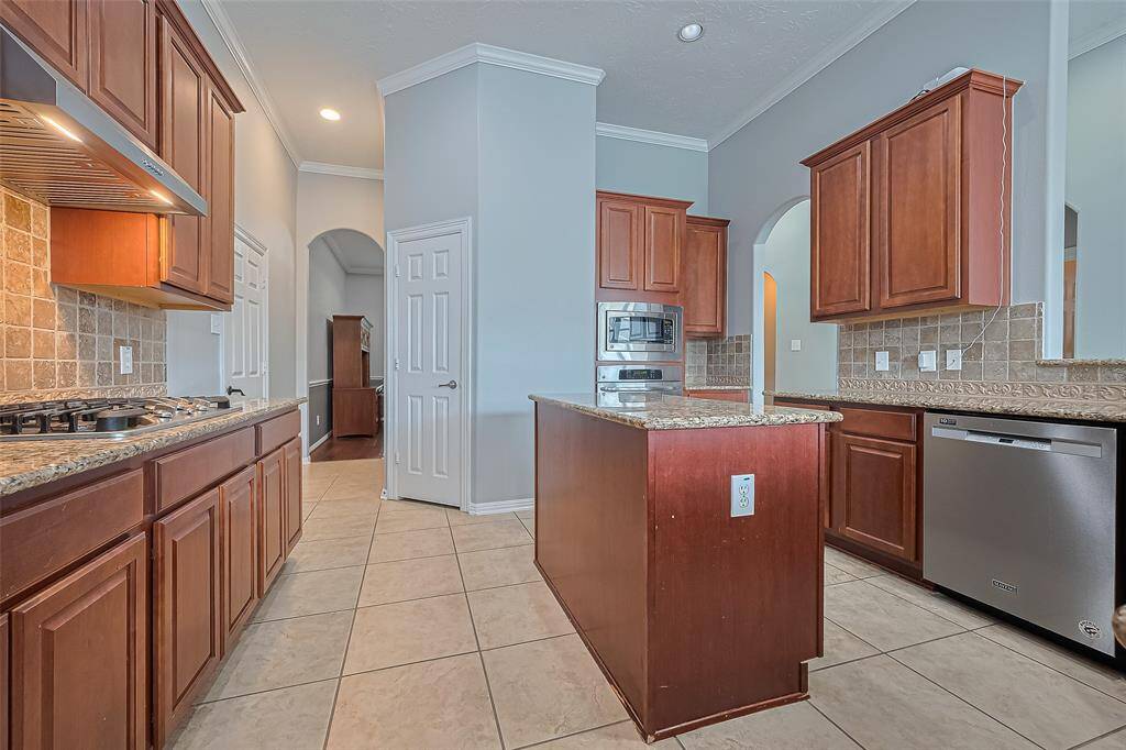 This kitchen features rich wooden cabinetry, granite countertops, and a modern stainless steel appliances. It includes a gas cooktop and built-in microwave, with tile flooring and a neutral color palette. An arched doorway adds a touch of elegance.