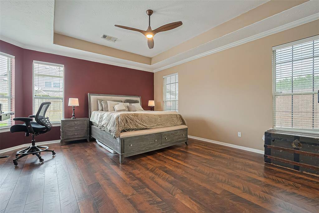 This spacious bedroom features hardwood floors, a tray ceiling with a modern fan, and ample natural light from multiple windows.
