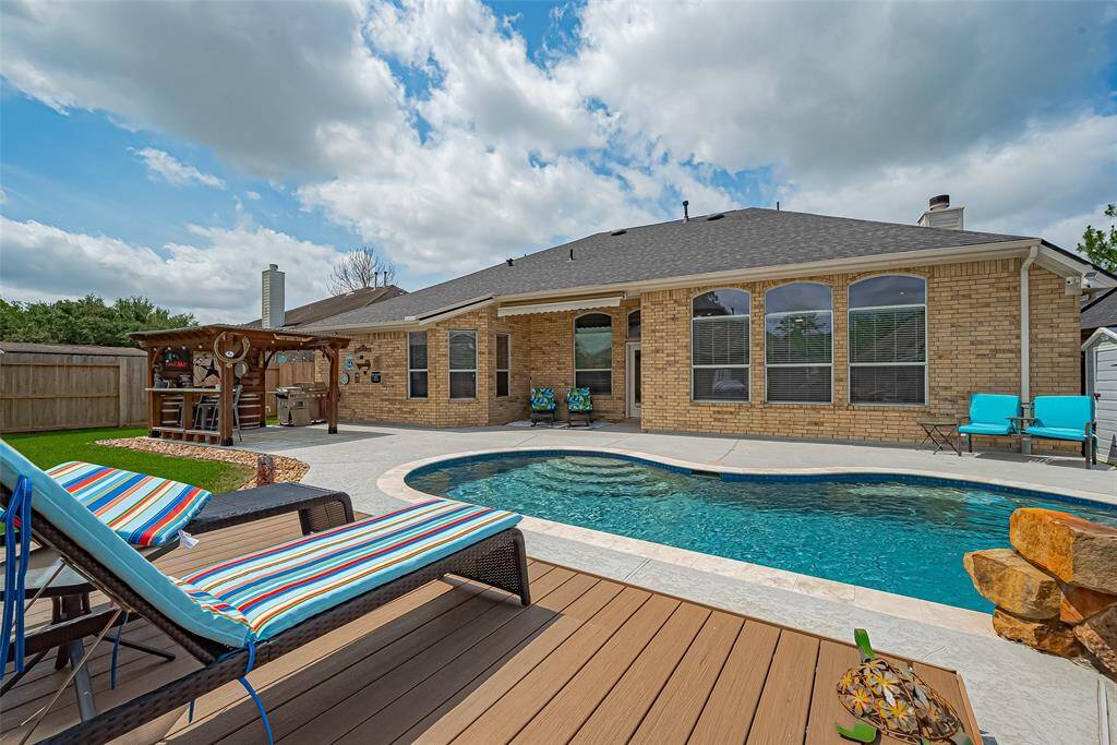 This photo showcases a spacious backyard with a recently added swimming pool and a pergola with an outdoor bar. Large windows from the home offer a welcoming and luxurious view of this beautiful outdoor living space.