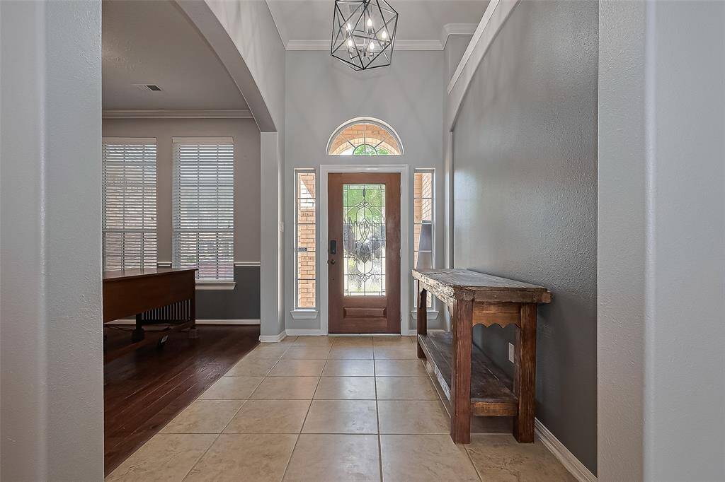 The entryway boasts a high ceiling with a modern chandelier, tile flooring, and a wooden front door with decorative glass. Side windows fill the space with natural light.