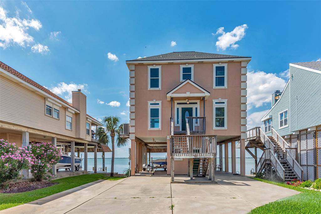 This elevated home features a spacious concrete drive, landscaped yard, and a covered ground-level area with walk-around access. The front entrance includes a set of stairs that leads to a covered entry before entering the main floor.