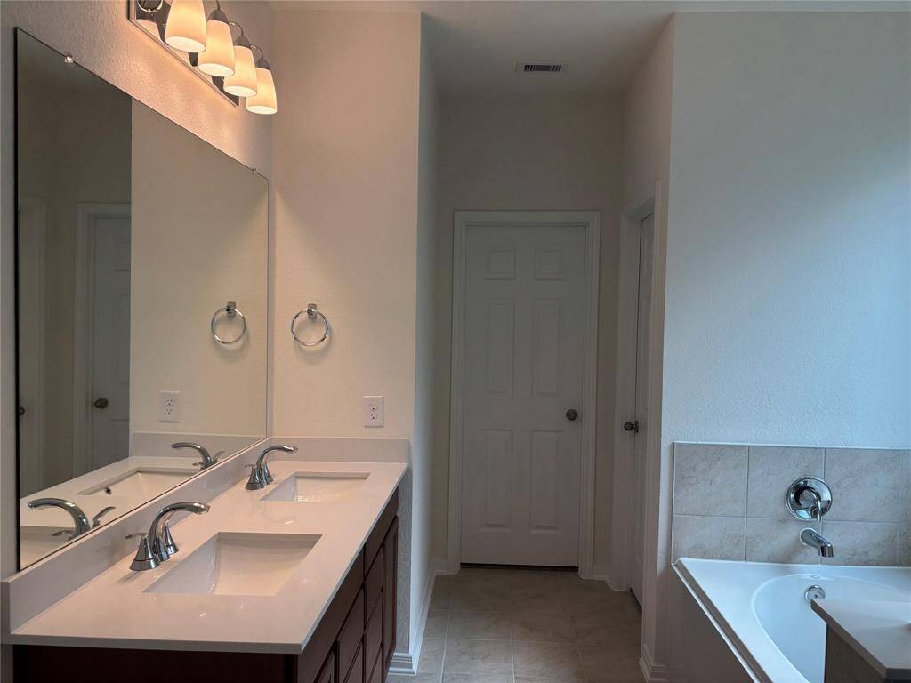 This primary bathroom features a double vanity with sleek faucets, a large mirror, and modern lighting. It includes a tiled bathtub, offering a clean and bright space.