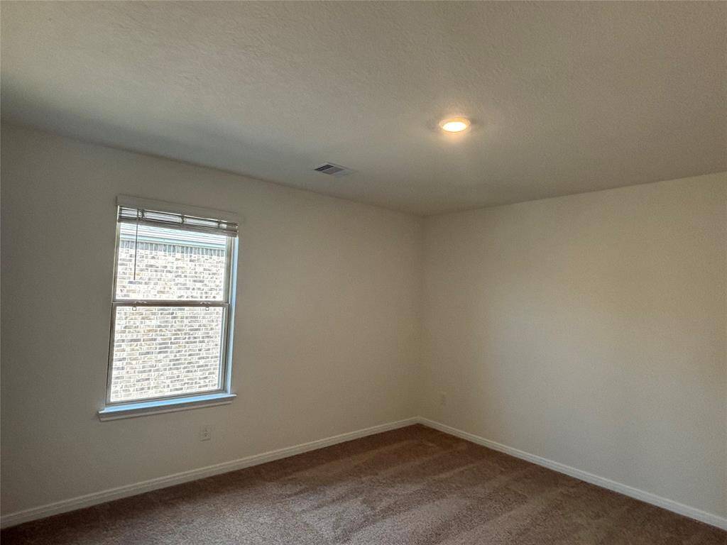 This is a second simple, unfurnished room with neutral-color walls and carpet. It features a single window allowing natural light and a ceiling light for illumination.