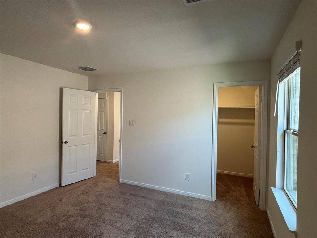 This photo shows a well-lit third bedroom with neutral walls and carpeted flooring. It features a window, a closet, and a door leading to a hallway.