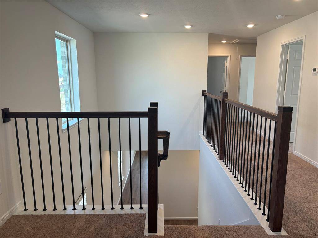 This photo shows a bright, carpeted upstairs hallway with modern railing, recessed lighting, and multiple doorways leading to other rooms. A window provides natural light, and the railing overlooks a staircase.