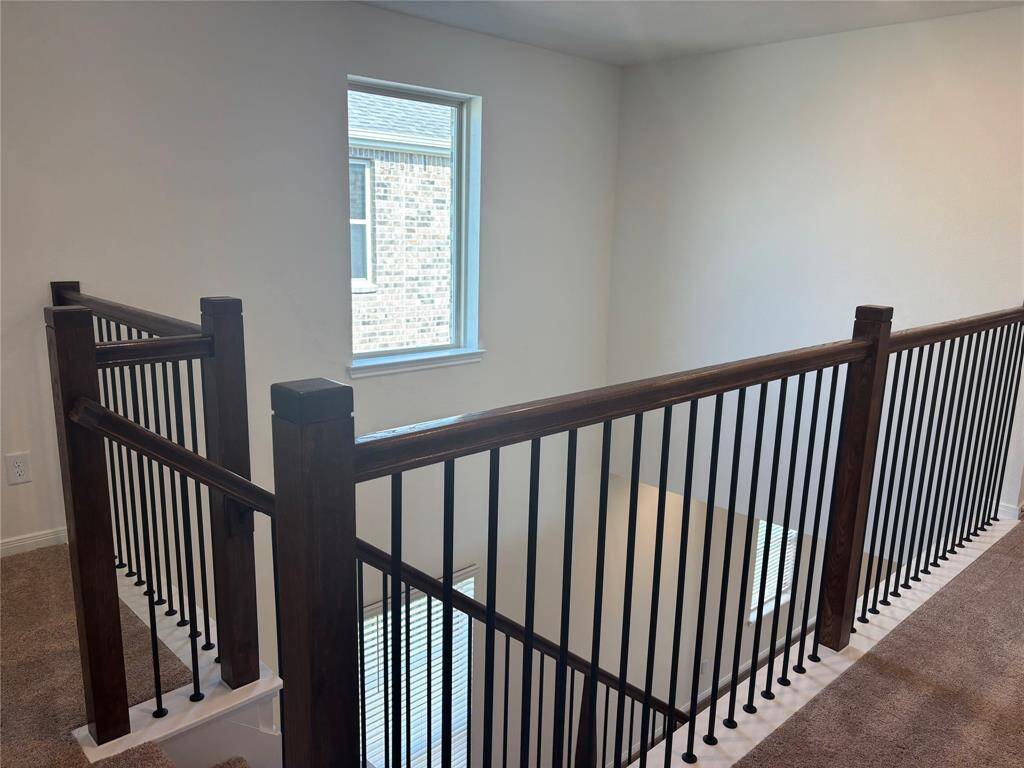 This photo shows a carpeted upper landing with a dark wood railing and black balusters. A window allows natural light, and you can see a view of brickwork outside. The area overlooks a lower level.