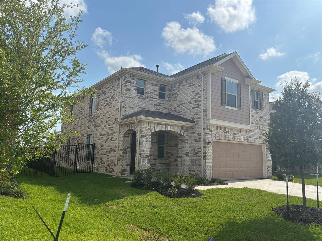 This is a modern, two-story brick home with a two-car garage and a neatly landscaped front yard. It features a charming front entrance with an arched doorway and is situated on a well-kept lawn under a clear blue sky.