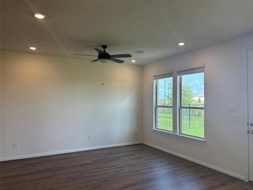 Bright, spacious room with wood flooring, recessed lighting, and a ceiling fan. Large windows offer natural light and views of a fenced backyard.