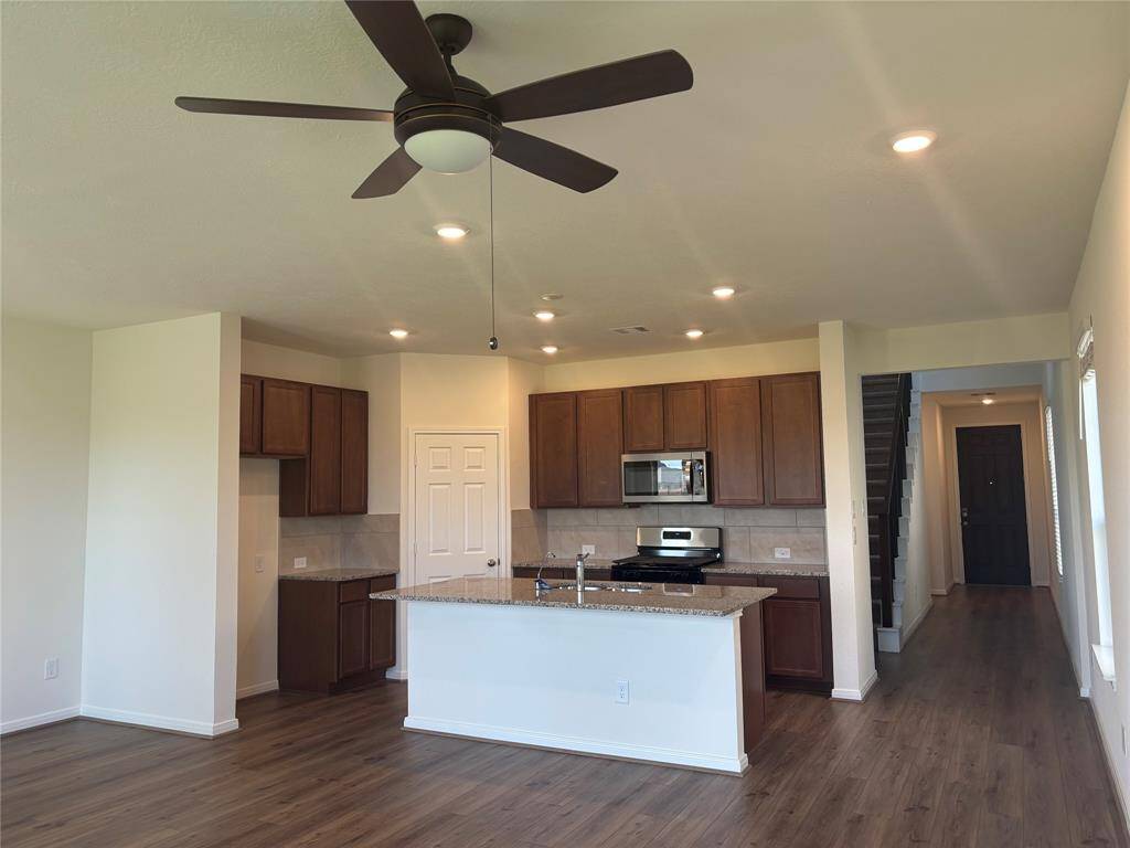 This bright and modern open-concept kitchen features wooden cabinets, a central island with granite countertops, and stainless steel appliances. The space is complemented by recessed lighting, wood flooring, and a ceiling fan, leading to a staircase and an entryway.